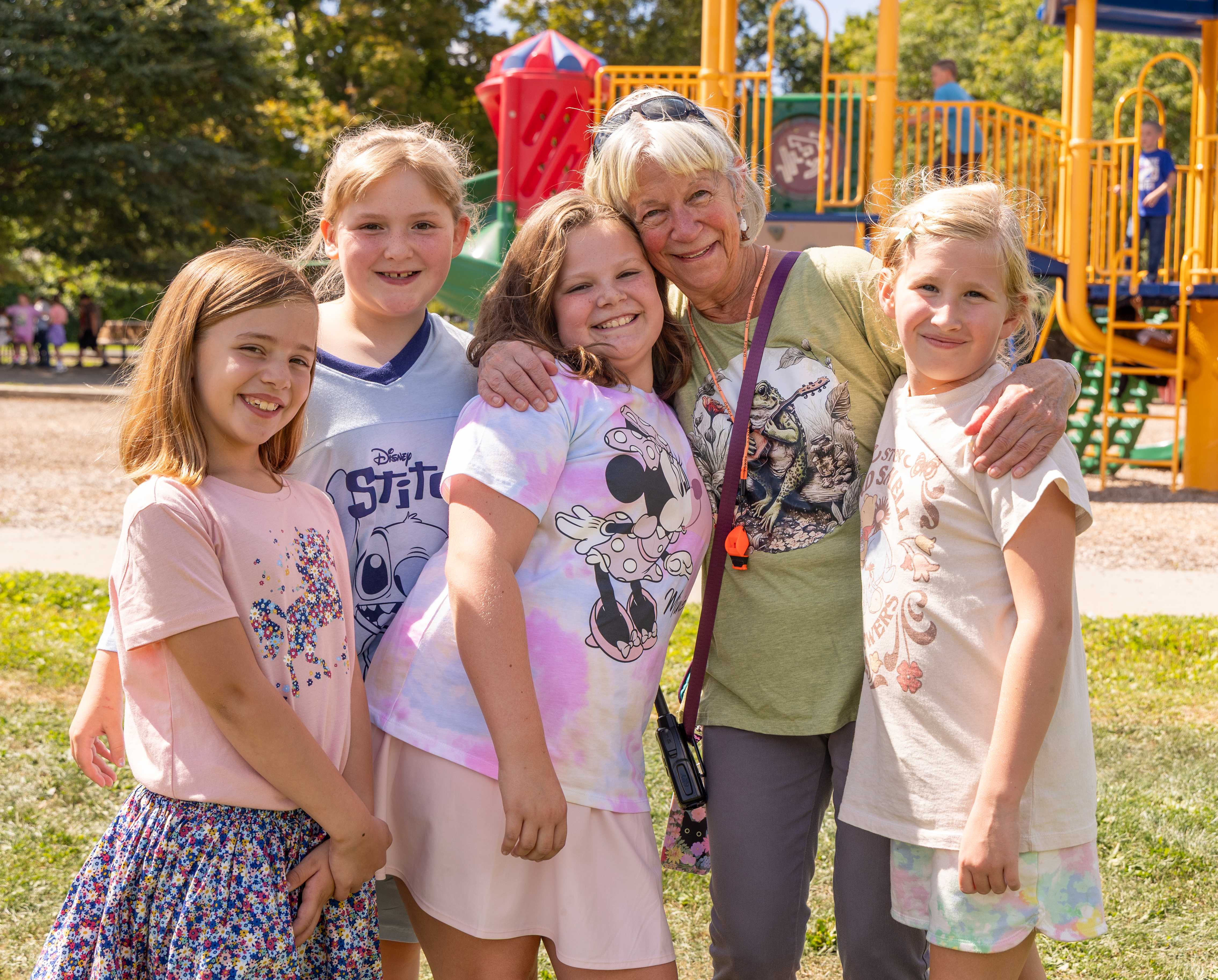 Ms. Patty poses with 4 young girls in front of the playground at recess.